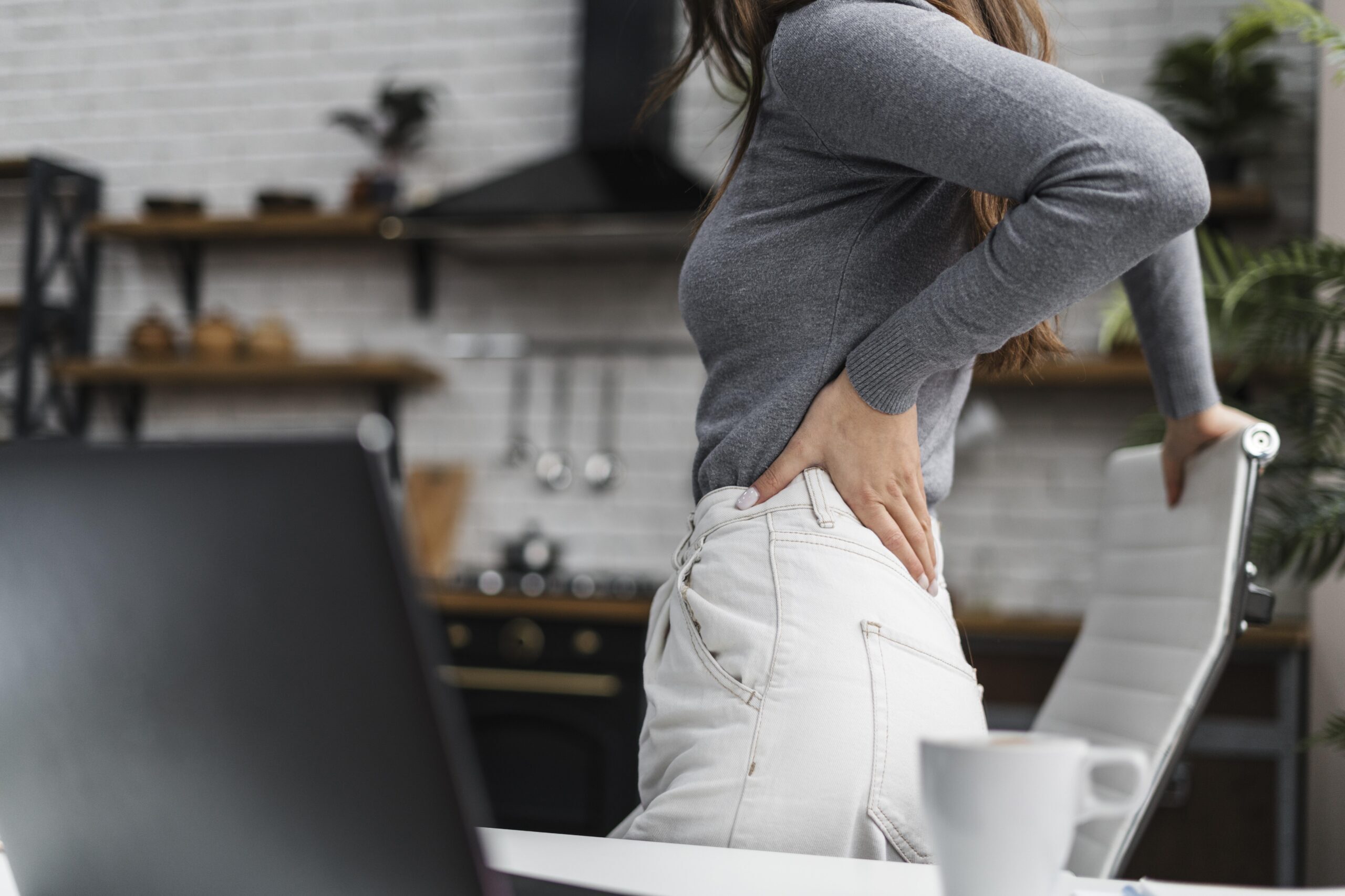 Mujer de pie junto a un escritorio se lleva la mano a la zona lumbar por dolor de espalda